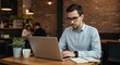 © FAWF STUDIO - A young man in his 30s, wearing glasses and a light blue dress shirt, is working intently on a silver ultra-thin laptop in a bright, modern coffee shop. In the background are exposed brick walls, plan