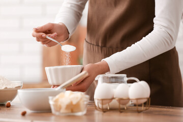 Wall Mural - woman adding sugar into bowl for preparing chocolate brownie at table in kitchen. closeup