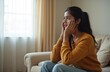 © Viktor - Pensive young Indian woman sitting on couch at home. Portrait of sad female in living room with thoughtful pensive expression, touching face. Mental health, depression, stress concept.