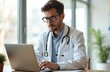 © Viktor - Young doctor in white coat working on laptop in office. Serious male gp consults patient online. Physician writing medical report or email. Healthcare worker is focused on work with stethoscope.