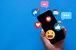 © Pakorn - Hand holding smartphone with social media notifications, likes, hearts, and emoji icons on vibrant blue background. Concept of online interaction, social network, and digital communication.