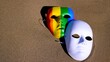 © Canary4stock - Two masks, one white and one painted in rainbow colors, resting on sand, symbolize the unveiling of true identity and the celebration of diversity during pride month