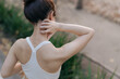 © Jirawatfoto - Asian woman experiencing neck pain after exercise, wearing white sports bra, standing outdoors in park with greenery in background, expressing discomfort