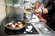 © pavel siamionov - A chef uses tongs to flip chunks of meat sizzling in a frying pan on a stovetop. The busy kitchen environment is lively, showcasing the art of cooking in progress.