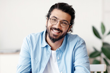 Poster - Portrait Of Happy Western Guy In Eyeglasses Sitting At Desk In Home Office, Smiling At Camera, Enjoying Remote Work, Cheerful Indian Freelancer Man Posing While Working With Laptop Indoors, Free Space