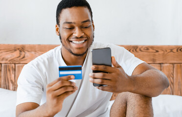 Poster - African American Man Using Mobile Banking Holding Smartphone And Credit Card Sitting In Bed At Home On Weekend. Online Shopping Application, E-Commerce Concept. Selective Focus