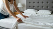 © Vasiliy - Woman making bed with white sheets and gray headboard in bright bedroom. Daily housekeeping routine maintaining clean sleeping environment and creating comfortable resting space