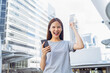 © sitthiphong - Cheerful young Asian woman holding smartphone with fists clenched celebrating victory expressing success in front of modern business building