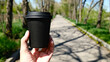 © Ployker - Person holding black paper coffee cup with lid against blurred outdoor park background, showcasing takeaway beverage