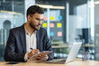 © Liubomir - A focused businessman, clad in a suit, meticulously takes notes while seated at a desk, utilizing a laptop in a modern office setting.