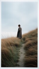 Poster - A solitary figure dressed in a long coat stands on a gentle slope, surrounded by tall, golden grass. The sky is a soft gray, creating a serene, reflective atmosphere