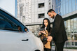 © wedmoments.stock - Business people man and woman with laptop discussing business project near electric car standing on charging. The concept of modern and environmentally friendly transportation and business travel.
