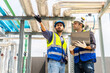 © TeTe Song - Collaboration, Two young construction workers discuss plans and use laptop computer while reviewing blueprints on a rooftop. They wear safety gear and oversee intricate piping systems in factory