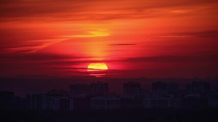 Wall Mural - Silhouette of city buildings against a red and orange sunset sky, time lapse. Urban skyline at dusk with large glowing sun. Evening cityscape.