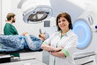 © wedmoments.stock - A confident medical professional looks into the camera in the background of a patient preparing for radiation therapy on a linear accelerator, looking confidently into the camera.