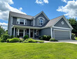 © TheseusStock - Photograph of an American-style two-story house with gray exterior walls, white trim and windows, set against the backdrop of lush green grass in the front yard, with a blue sky above.