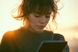 © Anjali - Young woman with windblown hair uses tablet at sunset, peaceful and serene moment.