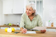 © JackF - Smiling elderly woman standing near countertop at kitchen at home, reading papers