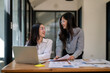 © Wasana - Two women are sitting at a desk with a laptop and papers in front of them