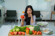 © MINAE - Young asian woman is sitting at a white table in her kitchen, holding a red bell pepper and looking pensive as she prepares a healthy salad with a variety of fresh vegetables