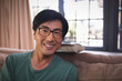 © WavebreakMediaMicro - Middle-aged Asian man sitting on beige sofa in bright living room, with eyeglasses and bag