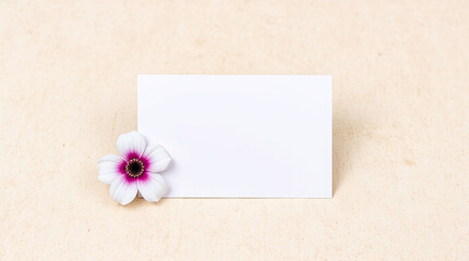 Poster - Blank white card rests beside a delicate flower. Clean background. Simple flower with purple accents. Ready for message. Nature art. Flat lay on tan textured surface. Gentle tones.