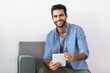 © wavebreak3 - Young adult Indian man sitting on modern gray couch in living room using white tablet, smiling