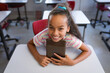 © WavebreakMediaMicro - Female child leaning forward on white desk in elementary classroom with red chairs, holding tablet