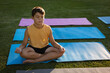 © WavebreakMediaMicro - Young boy meditating cross-legged on blue yoga mat on grass lawn, with pink and blue mats