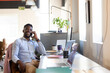 © Wavebreak Media - African American man sitting at desk in modern office, holding smartphone and looking at laptops