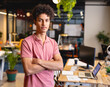 © Wavebreak Media - Young adult man standing with arms crossed in modern open plan office with laptop and plants