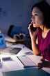 © Wavebreak Media - Asian Chinese woman talking on smartphone at dimly lit office, surrounded by laptop and keyboard
