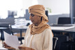 © Wavebreak Media - Mid-adult Indian woman with beige headwrap studying document at open-plan office, holding pencil