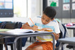 © Wavebreak Media - School age child writing with pencil in notebook at school desk in classroom, focusing on learning