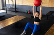 © Wavebreak Media - Woman performing floor exercise in indoor gym, holding red medicine ball near kettlebells and rings