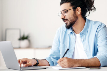 Poster - Young indian man entrepreneur using laptop and taking notes while working in modern office, sitting at desk and looking at computer screen, wester freelancer guy writing in notepad, copy space