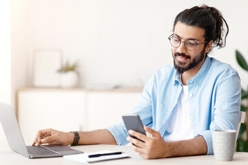 Poster - Millennial Arab Freelancer Guy Using Smartphone While Working On Laptop At Desk In Home Office, Browsing New Application Or Messaging With Friends, Enjoying Remote Job Opportunities, Copy Space