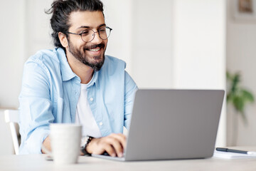 Poster - Young Indian Male Writer Using Laptop Computer, Sitting At Desk In Home Office, Handsome Western Man In Eyeglasses Typing On Keyboard And Looking At Screen, Enjoying Working Remotely, Selective Focus