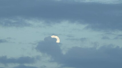 Wall Mural - full moon emerging from behind clouds with an early evening blue sky in the background.