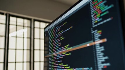 Aufkleber - Colorful programming code scrolls on a computer monitor, set against a blurred office background with a window and soft lighting, suggesting a focused work environment