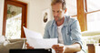 © peopleimages.com - Documents, reading and man in kitchen planning for debt, mortgage or tax payments in home. Paperwork, digital technology and male person with credit application, savings or monthly budget in house.