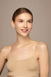© Beauty Agent Studio - Sincare beauty portrait of a young woman with smooth skin and slicked-back blonde hair smiling gently, wearing beige underwear top, captured in soft studio light on neutral background.