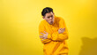 © Daniel - Cold young Asian man in yellow hoodie hugging himself tightly with scrunched face, as if shivering, standing against solid yellow background. Studio expression for chill, winter, or discomfort
