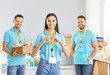 © Studio Romantic - Portrait of joyful caring volunteer team standing in humanitarian aid center together. Smiling young woman showing heart gesture with her hands with cheerful male colleagues holding food donations.