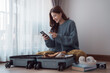 © Tj - Happy young woman packing clothes in suitcase and using smartphone, checking her travel itinerary or online booking information while preparing for vacation trip