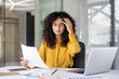© Liubomir - A concerned woman reviews documents in her office, appearing stressed while working on her laptop and analyzing papers at her desk.