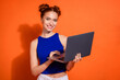 © deagreez - Smiling young woman with red hair holds a laptop while standing in front of an orange background, promoting a cheerful and modern lifestyle