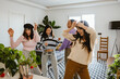 © Drobot Dean - Four diverse women wearing casual clothes, Asian, White, and Latino dance and laugh together in a sunlit living room. They enjoy a fun, spontaneous party moment surrounded by plants.