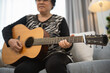 © WMSTUDIO - Older woman playing acoustic guitar at home showcasing passion for music and creativity, with comfortable living space as background setting