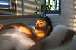 © Wavebreak Media - Reclining woman soaking in bathtub, with bubbles, plant and window blinds casting sunlight shadows